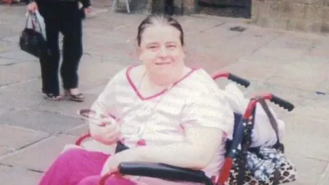 Family handout Angela Lloyd sits in her wheelchair and smiles at the camera. She is wearing pink clothes and has brown hair pulled back across her head.