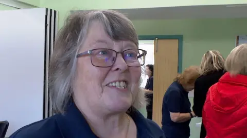 A woman stood in a room. She is wearing glasses and is wearing a blue polo shirt. She is smiling. There are people to the right of the woman in the background with their backs to the camera.