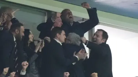 Prince William pictured with his arms in the air celebrating with people around him in the football stands also clapping, smiling and hugging.
