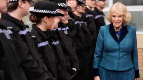 PA Media Queen Camilla smiles as she walks past a line of police officers in uniform. She has blonde hair flicked out to the sides and is wearing a blue tartan suit.