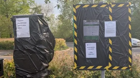 A parking machine covered in black tape, with a sign on it, next to a sign also covered in black plastic, with a number of notices on it. A car is behind the sign, by buses and trees. 