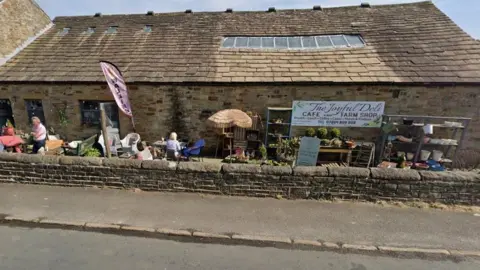 A cafe and farm shop set back behind a dry stone wall. Furniture, plants and plant pots are all in the courtyard outside. Some people are sitting on the chairs at tables on what looks like a sunny day.