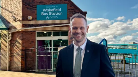 LDRS A man wearing a black jacket, white shirt and grey tie standing in front of the entrance to a bus station