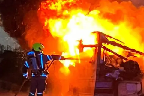Supplied Jenny Olley fighting a fire in full gear, including breathing apparatus and a yellow helmet. She is facing away from the camera, using a hose to spray water on a fire in a vehicle.