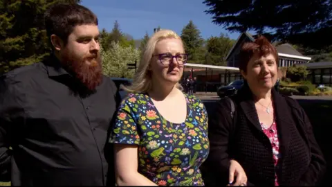 Three people are standing in front of a crematorium building which is surrounded by trees. On the left is a man in a black shirt with brown hair and a long beard looking to the side. In the middle is a woman with blonde hair, purple-rimmed glasses and a flowery top. She has her arm linked with an older woman next to her who is smiling and has short brown hair, and is wearing a brown cardigan and pink flowery top.