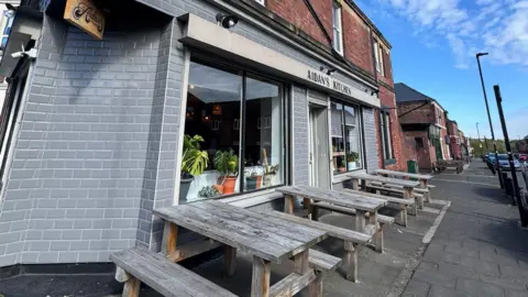 AIDAN JACKSON Aidan's Kitchen in Sandyford a cafe with outdoor seating. It has a grey exterior with a sign which reads "Aidan's Kichen" and there are four wooden benches in total outside of the street. 