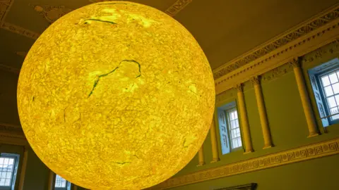The Helios sculpture during a previous display. It is a large sphere shape and has been lit up in a yellow colour.