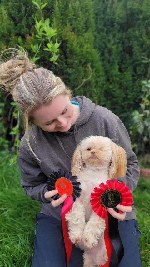 Lauraine Maddox A woman, looking down at a shih-tsu dog standing on it's hind legs. She is wearing a grey hoodie and blue trousers and has her hair in a bun. She is sat on the grass. She is holding two celebratory ribbons which are black and red.