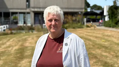 The image shows a woman wearing a burgundy t-shirt under a white blazer looks into the camera while standing on a large garden of grass in the sunshine. 