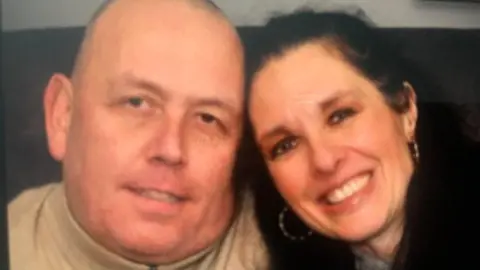 A close-up shot of a man and woman sitting together and smiling at the camera. The man has a shaved head and is wearing a beige tracksuit top. The woman has dark brown hair and is wearing large hooped earrings.