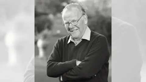 A black and white picture of a man with glasses and thinning hair. He is smiling and crossing his arms. He is wearing a dark jumper over a shirt.