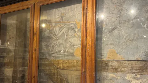 Katy Lewis/BBC A mural in a bad state of repair on a wall, with glass covering it. The picture is grey and rather faded.