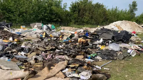 Gravesham Borough Council Large piles of dumped and burnt waste spread across open land in Northfleet, including household rubbish, carpets, timber, plastic containers, blackened debris and construction materials, with trees and fencing in the background.