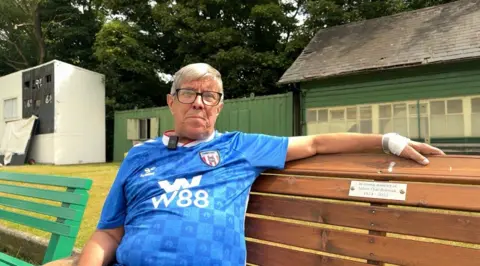 Molly Smith / BBC Alan is sitting on a brown bench which is dedicated to someone's memory but that plaque is not legible. He is wearing a blue football top, his arm is outstretched along the top of the bench. He has grey hair, glasses and is smiling slightly. 