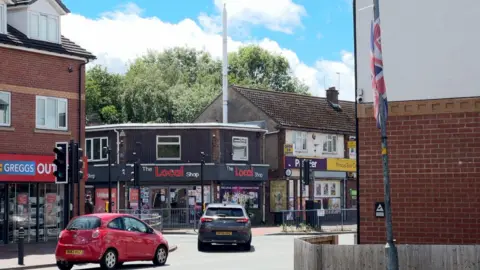 Another photo of Princes End. This photo shows two cars driving towards a traffic light junction with retail businesses on the left hand side.