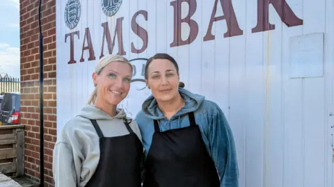 Candice and Kerry stand outside, shoulder to shoulder, smiling under a mural reading "Tams Bar". They're wearing grey and blue hoodies respectively under plain black aprons. 