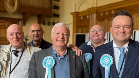 New councillors from Reform stand together and smile at the camera. The five men are wearing blue party rosettes on their suits and casual clothing.