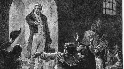 Getty Images Black and white drawing of a man in light-coloured clothes and a black jacket. He is standing in front of a group of prisoners, some of whom have their hands raised. He is pointing in their direction while holding a top hat with the other hand.