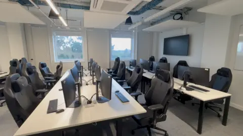 Josh Lovatt/BBC A small classroom has rows of tables with desktop computers and game chairs. There are four computers on each row and there is a monitor on the wall on the right.