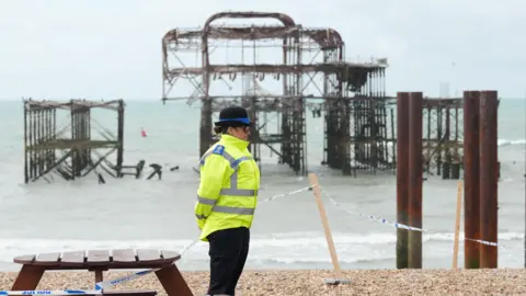 A police officer wearing a yellow hi-vis jacket stood on a pebbly beach inside a cordon. The remains of a pier sit in the sea in the background.