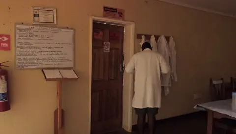 TRACE, Simon Dures A man in a white coat stands to the right of the picture with his back to the camera.  He is in a dark corner of a room with walls painted in a light yellow colour.  The room has a noticeboard on the wall to his left beyond a door.  At the top of the noticeboard a sign reads "Lab actions and Projects".