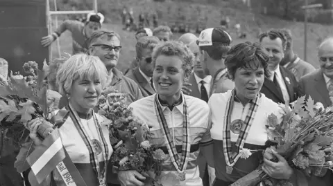 Leeds City Council A black and white photograph of three women in cycling gear. They have medals around their necks and are holding flowers. Behind them stands a group of men in suits.