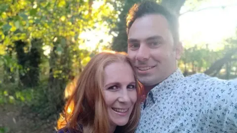 A man and woman posing together for a photo. A row of trees with green leaves are in the background.