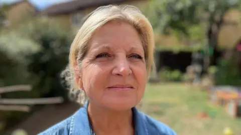 DJ McLaren/BBC A woman in a garden on a sunny day. She is smiling at the camera. She has blonde hair that has been tied back. She is wearing a denim top. The garden behind her has been blurred.