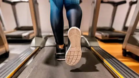 A person wearing blue leggings and black trainers with white soles on a running machine.