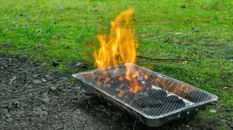 A disposable barbecue straddles a soily patch and grass as it burns.