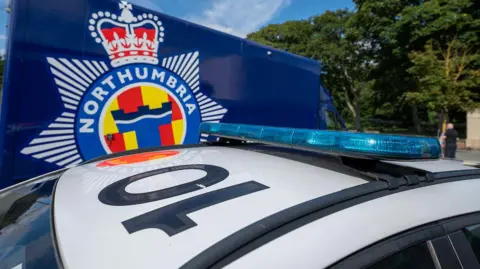 The white roof of a police car with the number 10 written in black lettering on it. Roof bars and a bar holding the blue light strip are black. A very small amount of the windows is visible and the car is parked in front of a royal blue van with a large blue, yellow, red and white, star-shaped Northumbria Police logo on the side.