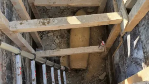 Ben Remfrey A deep, rectangular excavation site reinforced with wooden planks. A ladder leans against one side, leading to the bottom where a rusted cylindrical object is partially buried in dirt, surrounded by rocks and debris.