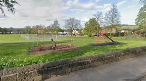 Google Menston Recreation Ground on a sunny day. Two sets of swings and a slide can be seen on a meadow. In the distance, rows of houses are dotted along the road.