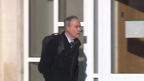 James Rouse has short grey hair and is wearing a shirt, tie and black coat. He is walking towards Portsmouth Crown Court.