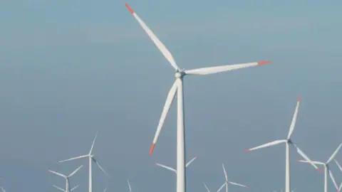 Offshore wind farm. there are about eight white wind turbines with orange on the tips. The background a blue sky.