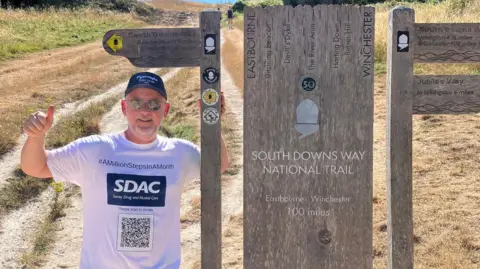 A man in a white T-shirt, sunglasses and a black cap gives the camera a thumbs up as he stands beside the South Downs Way National Trail sign