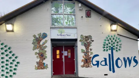 The front entrance of a single‑storey community-style building with a peaked roof. The exterior walls are painted white, and the entrance features two bright red double doors in the centre. Above the doors is a sign that reads “Gallows Close Centre”, accompanied by a tree-like logo. On both sides of the doors are colourful, decorative murals of trees.
