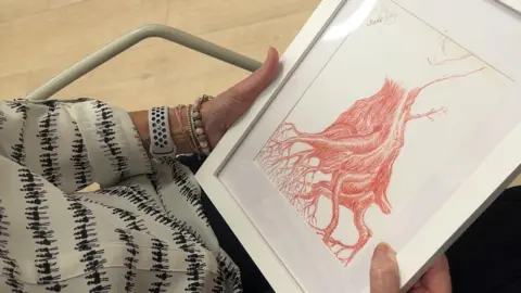 A woman's hands hold a drawing in a white frame. It depicts the trunk of a tree with long winding roots and has been sketched in a red pencil on a white background.
