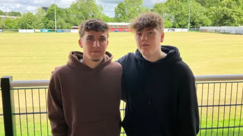 George Callie and Tom Gough stand in front of a football pitch. They are both wearing hoodies and have their arms wrapped around each other. 