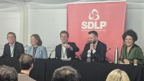 SDLP party members joined by Fleur Anderson sitting at a conference table with an SDLP poster behind them 