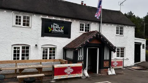 The pub is painted white with a black roof, sign and porch area. It has a Union Jack on top of the porch entrance and beer banner adverts either side of the entrance.