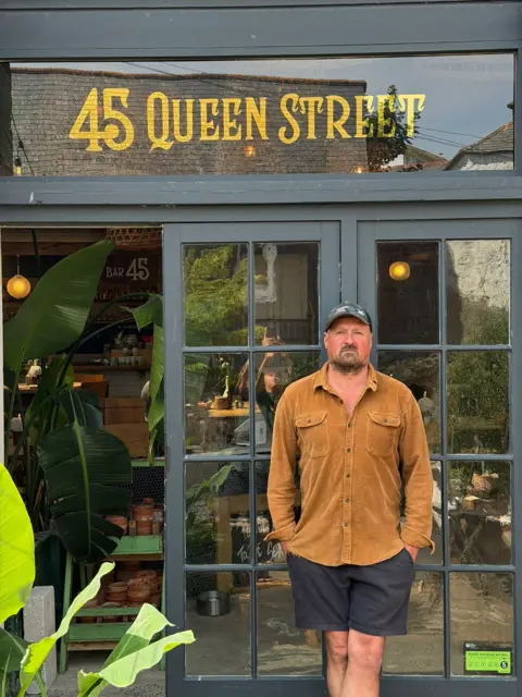 Sam Lamiroy Sam Lamiroy, a tall white man with a beard, stands outside 45 Queen Street, his bar and restaurant in Penzance, wearing black shorts, a brown shirt and a cap.