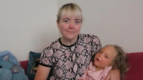 A woman in a pink alligator skin print t-shirt, with bleach blonde hair smiles at the camera, who has honey blonde hair, and looks at the camera. They are sitting on a red sofa with a blue stuffed toy next to them and a white wall behind. 