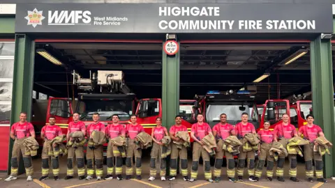 The male and female firefighters stand in a line at the entrance to their station with fire engines behind them. They are wearing pink T-shirts and their brown/black uniform trousers and are holding the top jackets.