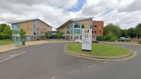Google Light coloured college building with a grey sign in front on a grass round verge, saying City of Wolverhampton College. A green bus stop is near the entrance as well as a red and white car.