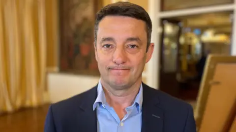 Boley Smillie - A man with black hair looking at the camera. He has a blue suit on and a blue button up shirt. 