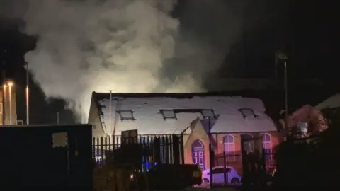 A large plume of smoke rises from a building with snow on its roof.