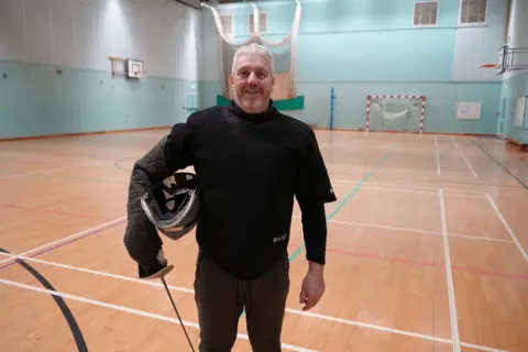 Sean Whitmore / BBC O técnico Paul está em um pavilhão esportivo com uma arma na mão direita e uma máscara de esgrima debaixo do braço.