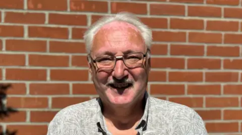 A man with glasses and grey hair smiling into the camera while standing in front of a brick wall.