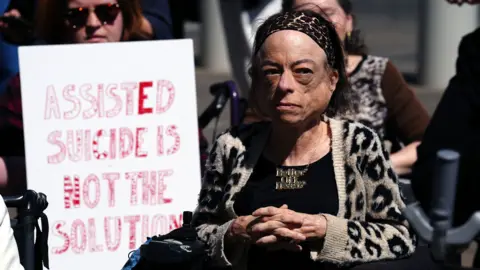 Actor Liz Carr, wearing a leopard print cardigan, black top and necklace. Next to her is a banner with the words 'Assisted suicide is not the solution'